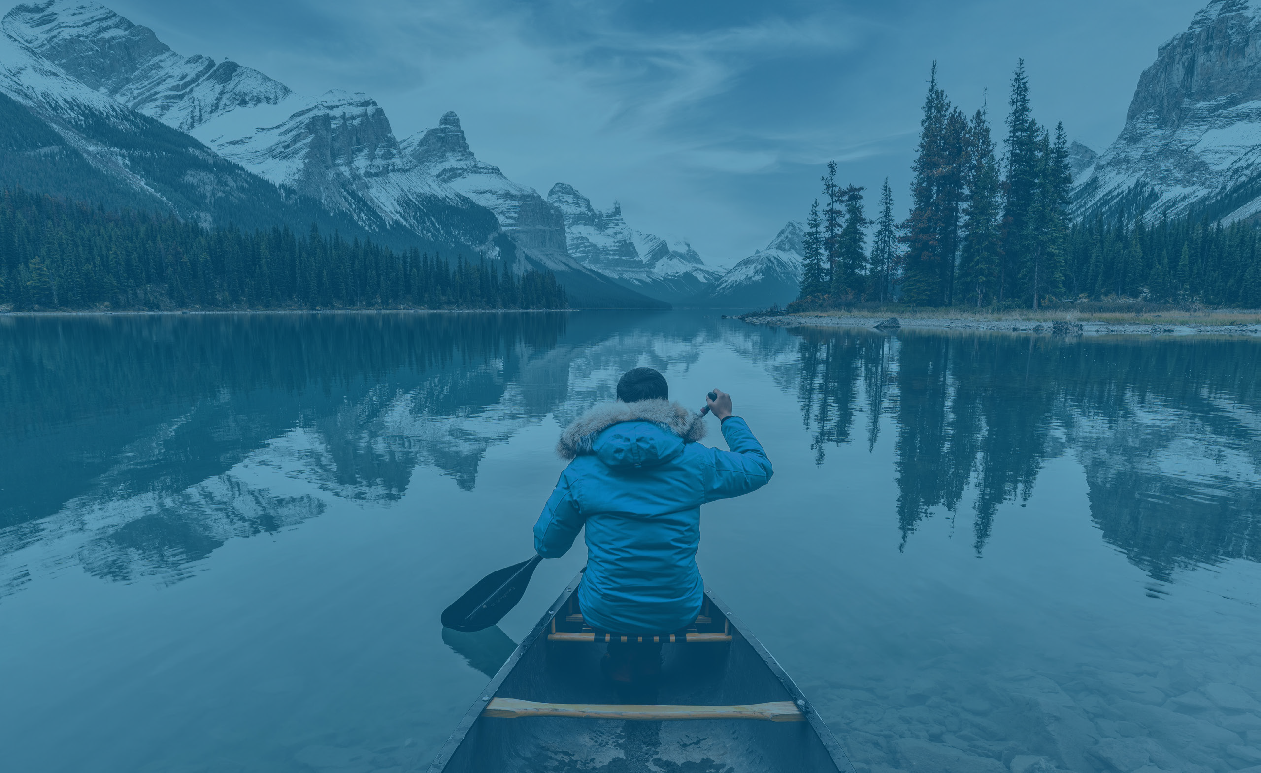 Une personne dans un canoë sur un lac tranquille regardant une forêt et des montagnes enneigées, vue de derrière
