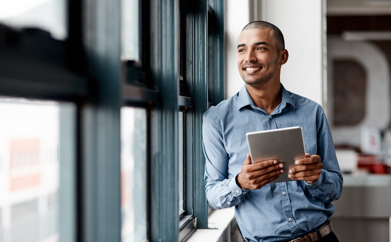 A man smiling and holding a laptop, looking out a window.