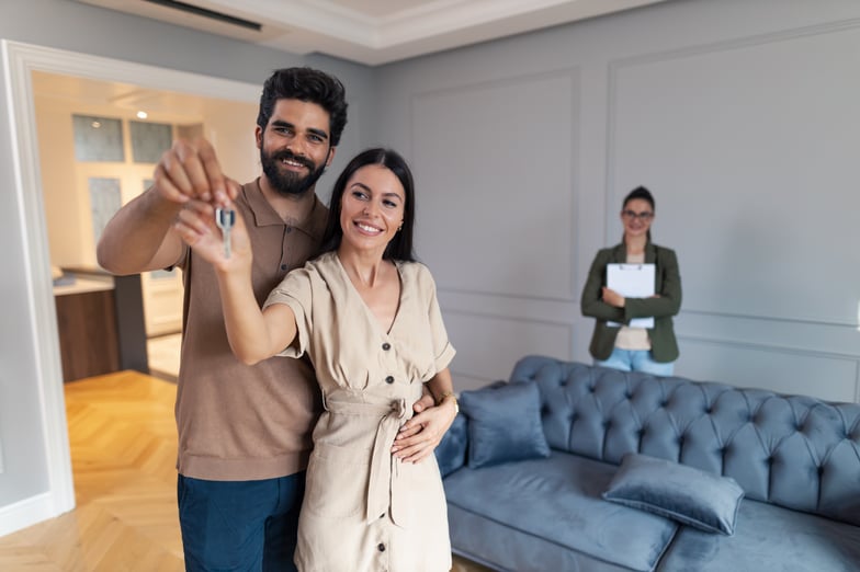 Image of a couple smiling holding keys in a new home with a realtor smiling in the background.