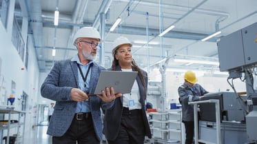 Photo d’une femme et d’un homme avec un ordinateur portable dans les mains, en train de marcher dans une usine. 