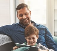 Un père et son fils assis sur le divan regardent ensemble une tablette électronique.